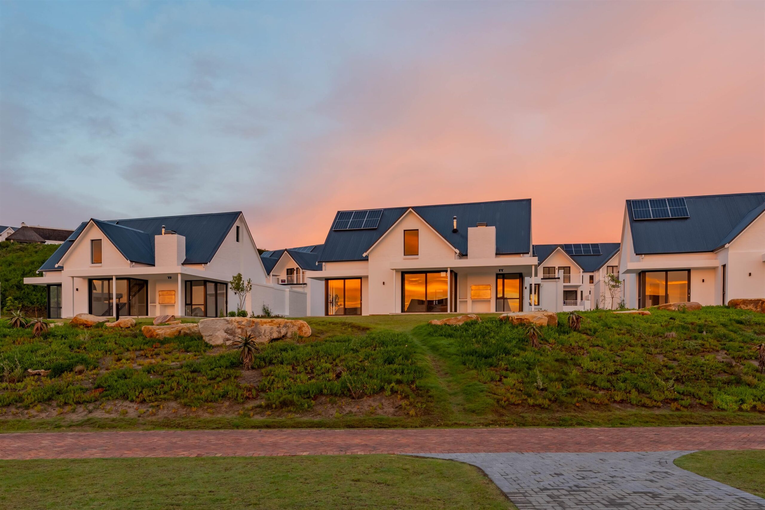 Modern white houses with solar panels on pitched roofs, glowing warmly at sunset on a landscaped hillside.