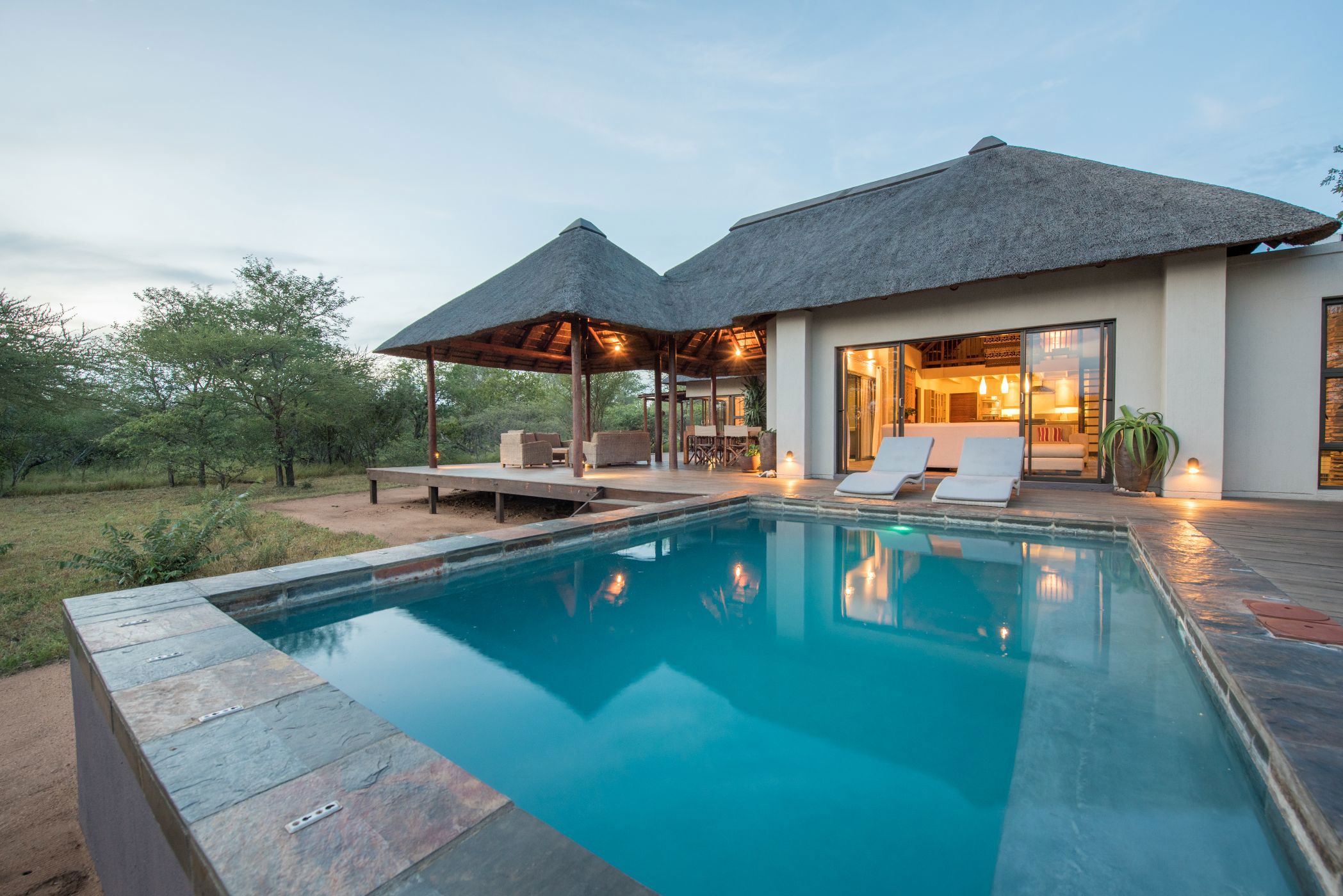 Thatched-roof house with wooden deck and swimming pool at dusk, surrounded by bushland.