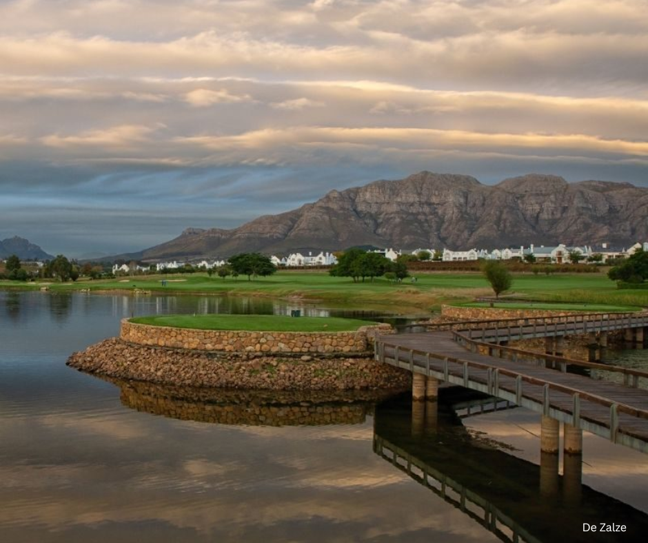 Wooden bridge over a calm lake on a golf course with mountains in the background.