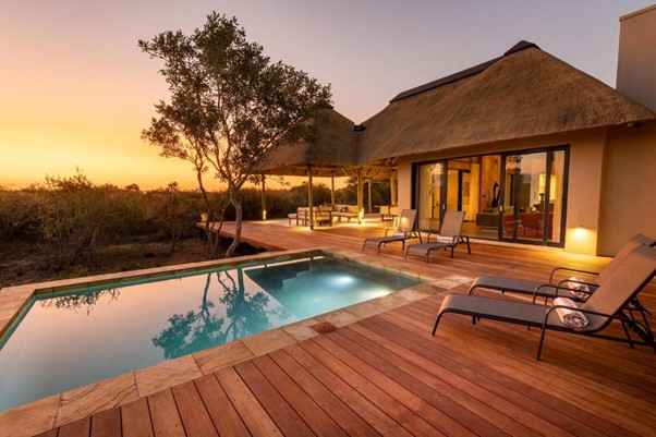 Thatched-roof house with pool and wooden deck at sunset.