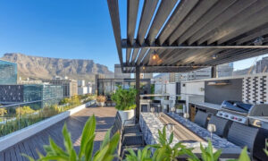 The rooftop terrace of the Onyx double penthouse in late afternoon light, with a steel pergola over a long dining table and built-in braai, timber decking running to a glass balustrade, and Table Mountain rising behind the Foreshore office towers