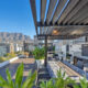 The rooftop terrace of the Onyx double penthouse in late afternoon light, with a steel pergola over a long dining table and built-in braai, timber decking running to a glass balustrade, and Table Mountain rising behind the Foreshore office towers
