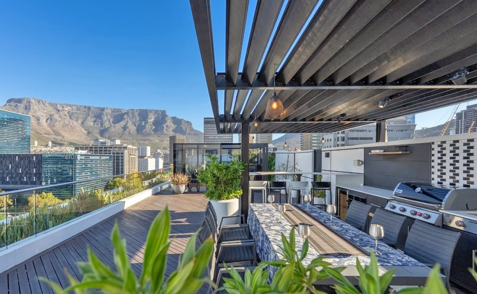 The rooftop terrace of the Onyx double penthouse in late afternoon light, with a steel pergola over a long dining table and built-in braai, timber decking running to a glass balustrade, and Table Mountain rising behind the Foreshore office towers