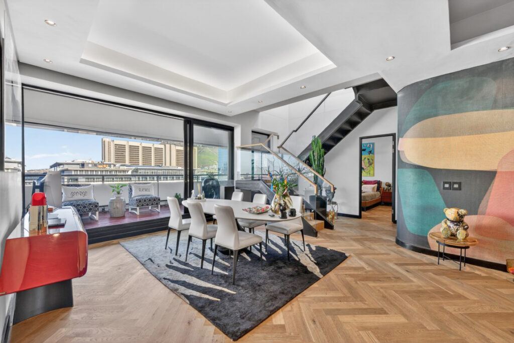 The penthouse dining and living level, herringbone oak floor, oval dining table, a black steel staircase rising to the roof level and glazed doors to the balcony