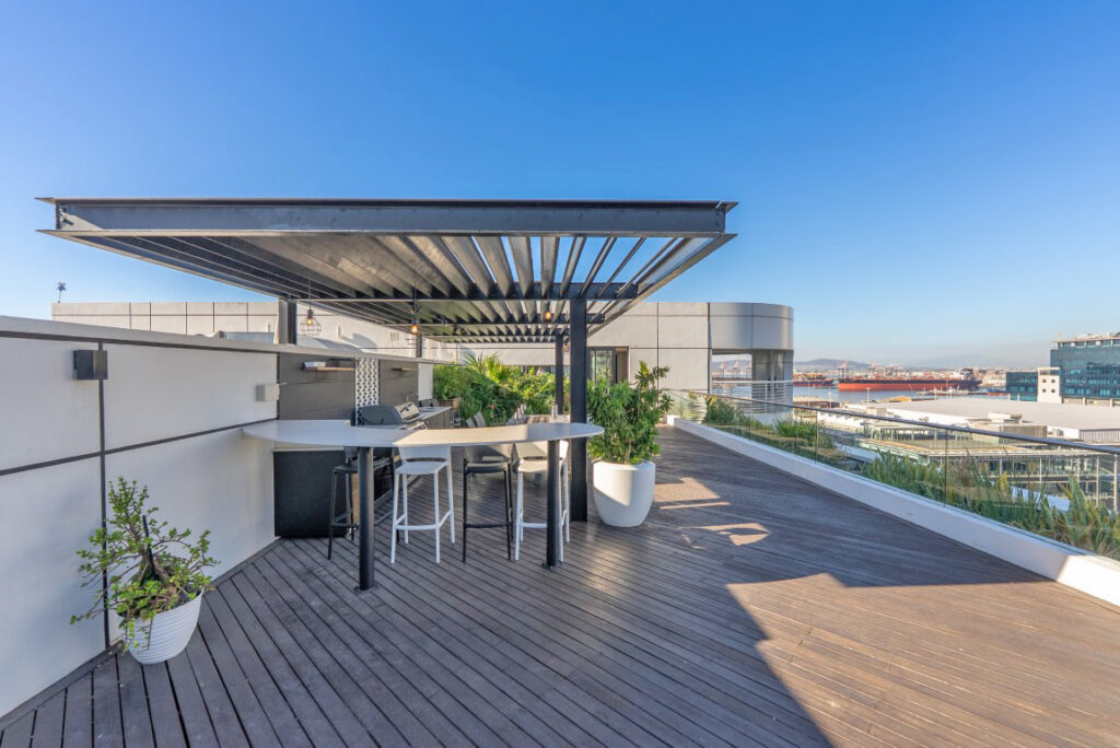 The upper roof terrace, a cantilevered steel pergola over a curved bar counter with a built-in gas braai, Table Bay visible beyond the glass balustrade