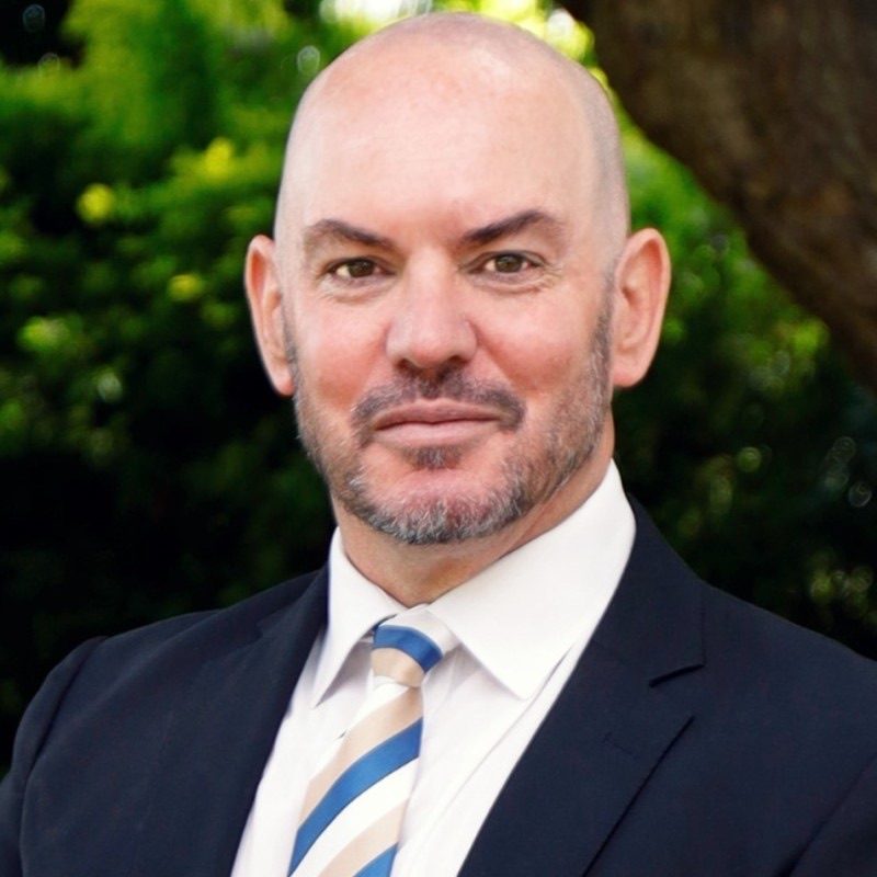 Greg Dart, Director at The High Street Auction Co, in a dark suit and striped tie against a garden background.