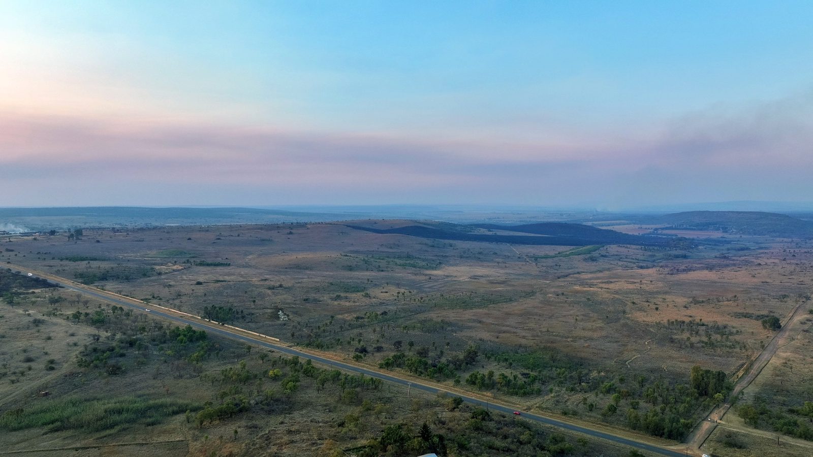 Aerial view of open highveld country at dusk, with the road crossing it and scattered buildings in the far distance.