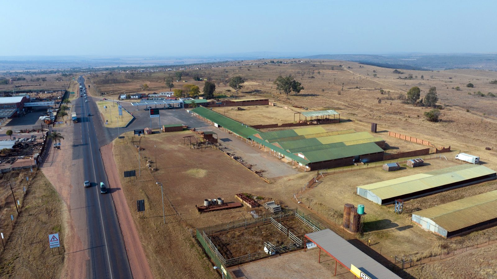 Aerial view along the R573 Moloto Road running straight to the horizon, with warehouse and retail buildings strung along its left-hand side and open veld on either side.